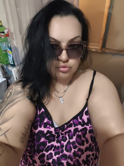 The image shows a close-up, selfie-style shot of a woman with dark hair, wearing sunglasses and a leopard-print camisole. She has a silver necklace with a cross pendant. Her left arm displays tattoos. The background is indistinct, with hints of curtains and possibly cleaning products on a shelf to the left, and a wooden surface to the right. There is no indication of the image being located in Komotini, Greece.