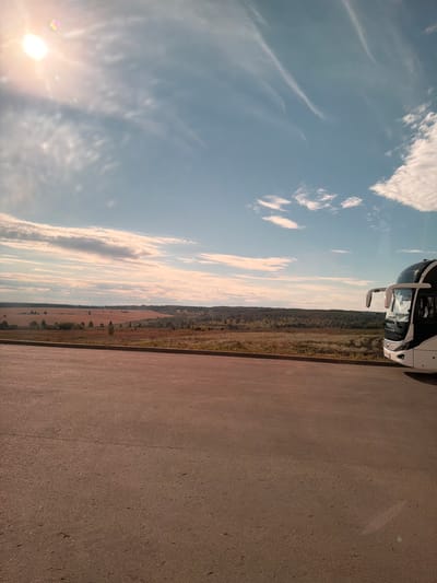 An outdoor, low-angle, medium shot captures a bus parked on an asphalt road in Priovrazhnoe, Russia. The bus, white with black accents, is positioned on the right side of the frame, facing left. Beyond the road, a vast, open landscape unfolds under a partly cloudy blue sky. In the mid-ground, fields of varying colors, possibly agricultural, stretch across the horizon, interspersed with scattered trees and a distant treeline. The sun is a bright, hazy orb in the upper left corner, casting a warm glow and creating lens flare. The foreground is dominated by the textured asphalt, showing a slight crack. The scene appears to be during daylight, with the sun's position suggesting it might be late afternoon.