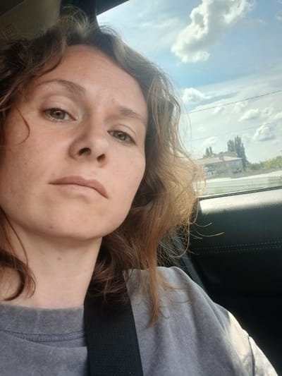 The image is a close-up, low-angle selfie of a person with light brown, wavy hair and fair skin, taken inside a vehicle. The individual is looking directly at the camera with a neutral expression. They are wearing a grey t-shirt and a black seatbelt is visible across their chest. The background through the car window shows a bright blue sky with fluffy white clouds, trees, and buildings, indicating an outdoor environment. A building with multiple stories and a peaked roof is visible, along with some foliage and power lines. The interior of the car is partially visible, including dark leather upholstery. The lighting suggests it is daytime. The location is identified as Latnaya, Russia.