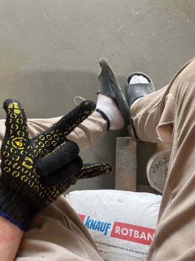 The image shows a close-up view of a person's legs and hand, likely in a construction or workshop setting. The person is wearing tan work pants, white socks, and dark gray slides. They are also wearing a black work glove with yellow patterns on their right hand, and are making a hand gesture. In the background, there is a white bag with "KNAUF ROTBAN" printed on it, and a container with "2002" written on it. The environment appears to be indoors with a concrete floor. The image was taken in Strumivka, Ukraine.