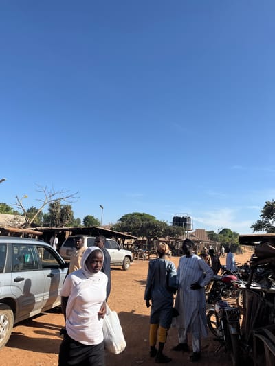 The scene depicts an outdoor public area in Vodni, Nigeria, under a clear blue sky, with unpaved reddish-brown ground. In the immediate foreground, a dark-skinned woman wearing a white headscarf, white t-shirt, and dark skirt walks towards the left, carrying a white plastic bag in her left hand. To her left, a silver Subaru Forester SUV is parked. Behind her, other individuals in traditional and contemporary attire are present near a parked white SUV. In the mid-ground, two men stand facing each other, one wearing a dark tunic over yellow pants and a light cap, the other a striped light blue and white tunic with his hands on his hips. To the right, several motorcycles are parked among rudimentary market stalls constructed from wood and corrugated metal. The background features scattered trees, some bare, and several utility poles equipped with solar-powered streetlights. A tall metal structure supporting two black water storage tanks is visible in the far right background. Multiple other individuals are discernible throughout the area, walking or standing. The scene is brightly lit by direct sunlight, casting distinct shadows.