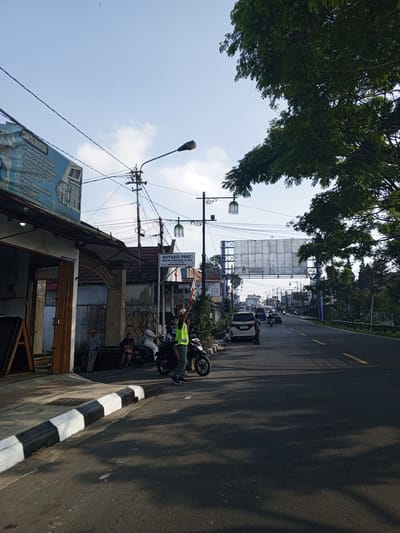The image displays a sunlit street scene in Weddagala, Sri Lanka, captured from a slightly elevated perspective looking down a two-lane asphalt road. A sidewalk with a black and white striped curb runs along the left side. Adjacent to the sidewalk, a single-story building features a large blue sign advertising construction services such as "Pembuatan Kusen" (frame making) and "Jendela Aluminium" (aluminum windows), with text presented in Indonesian. Further along the sidewalk, an ornate archway precedes a white sign for a notary and land deed official, "NOTARIS/PPAT ANGELINA LETUNGGANU, S.H., M.Kn.", also written in Indonesian.
Overhead, a dense network of utility wires spans between multiple utility poles, interspersed with ornate streetlights featuring multiple lanterns. In the mid-ground, a large, rectangular metal billboard structure, currently unbranded, bridges across the width of the road. Vehicles, including several motorcycles and a white van bearing the Indonesian license plate "B 2426 KJM", are visible on the road and parked along the shoulder.
In the foreground, a man wearing a bright yellow-green vest and dark pants stands on the sidewalk, holding a red-tipped baton, suggesting traffic management. To his left, two other men are seated on a parked motorcycle on the sidewalk. Long, distinct shadows from trees and unseen structures are cast across the asphalt road and sidewalk, indicating bright, directional sunlight. The upper right portion of the frame is filled with the dense green foliage of a large tree. The sky above is clear blue with a few scattered white clouds.