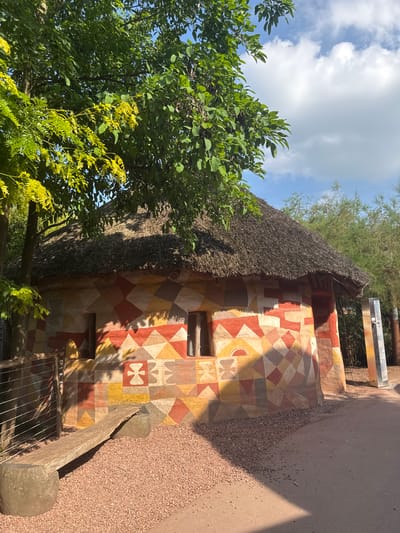 This is an outdoor, eye-level, medium shot of a brightly painted, round hut with a thatched roof, located in the Pairi Daiza zoo in Cambron-Casteau, Belgium. The hut's exterior walls are adorned with a geometric pattern of colorful rhombuses and triangles in shades of red, yellow, and beige, with some areas featuring stylized symbols. There are two small, dark windows on the side of the hut facing the camera. A wooden ramp leads up to an opening on the left side of the hut. In front of the hut is a rustic stone bench supported by two large, rounded stones, placed on a bed of reddish-brown gravel. A wire fence with wooden posts is visible to the left of the bench. Lush green trees with dappled sunlight filtering through their leaves partially obscure the top of the hut and extend into the background, where more trees and a bright blue sky with scattered white clouds are visible. A tall, grey metal structure stands to the right of the hut. The ground in front of the hut is a mixture of gravel and dirt path.