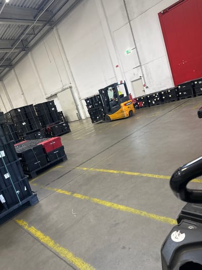 The image depicts the interior of a warehouse with a high ceiling. The floor is concrete with yellow lines painted to mark pathways. The scene is dominated by tall stacks of black plastic crates, some of which are filled with items. A few red crates are interspersed within the stacks.
In the foreground, a yellow forklift is being operated by a person wearing a yellow hard hat and a grey shirt. The forklift is positioned near the stacks of crates, suggesting ongoing operations like loading or unloading. The overall atmosphere appears to be one of active industrial work.
The warehouse has a red sectional door at the far end and a white wall with some electrical conduits and lighting fixtures. The ceiling is made of concrete panels, with exposed pipes and wiring running across it. The lighting suggests it is daytime, but the weather is not discernible as it is indoors.
Some visible text includes "Jungheinrich" on the forklift and "TX" on some of the crates. The location context is Schweitenkirchen, Germany. The image is taken from a high-angle perspective, looking down into the warehouse.