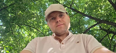 A man wearing a light-colored baseball cap and a light-pink polo shirt is photographed from a low angle looking upwards. The man has fair skin, light-colored eyes, and stubble on his face. He is positioned centrally in the frame, with his head and upper torso visible. The background is dominated by the lush green foliage of a tree, with sunlight dappling through the leaves, creating a bright and natural outdoor setting. There is no indication of the specific location within Collado Villalba, Spain, in the image itself.