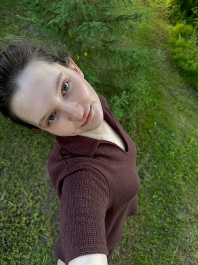 A high-angle, wide shot captures a young woman with fair skin and light blue eyes looking upwards at the camera. She has dark brown hair pulled back, with a few strands framing her face. She is wearing a long-sleeved, collared, brown knitted shirt. Her head is tilted to her left. The background consists of lush green grass and the branches of a pine tree. The scene appears to be outdoors, in a natural setting that could be located in or near Burdyata, Russia.