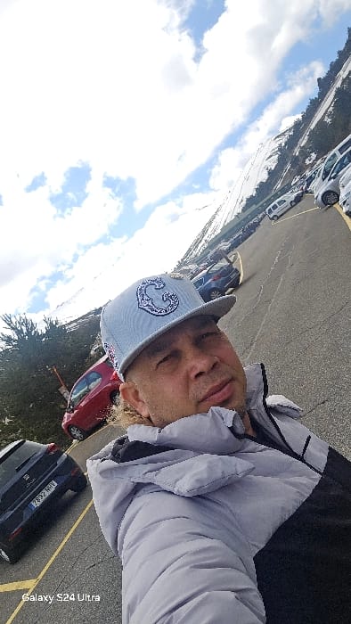 The image is a diagonal, low-angle selfie featuring an adult male in the foreground of a mountain parking lot located in Rascafría, Spain. The man, displaying light skin and stubble, wears a light blue baseball cap with a white stylized 'C' logo and a two-tone puffer jacket, light gray on the upper portion and dark gray/black on the lower. He looks directly at the camera with a neutral expression.
The background shows an asphalt parking lot with visible cracks and yellow line markings, containing multiple parked vehicles. A dark blue SEAT hatchback with the Spanish license plate '7892 HHV' is visible on the left, along with a red hatchback further back. Beyond the parking area, snow-covered mountains dotted with evergreen trees rise under a partially cloudy blue sky, indicating a bright, cool weather condition at a high-altitude setting.
