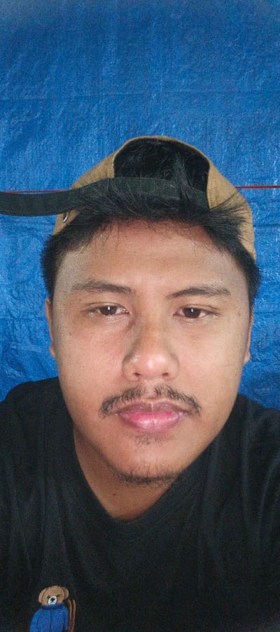 A close-up, waist-up, selfie-style photograph features a person wearing a black t-shirt and a tan baseball cap with the brim tilted back. The person has dark hair, eyebrows, and a mustache, with light stubble on their chin and jawline. Their eyes are dark brown and looking directly at the camera. The background is a textured blue tarp. The image was taken in Banding Agung, Indonesia.