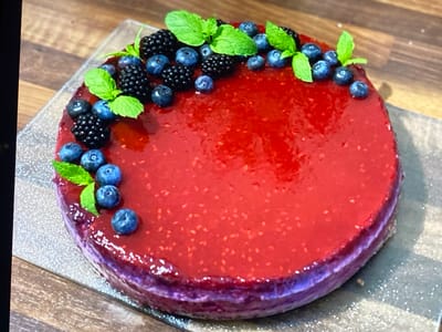 A round, layered cake topped with a vibrant red glaze and garnished with fresh blueberries, blackberries, and mint leaves sits on a silver serving platter. The cake has a dark purple base layer and appears to be a dessert. The platter is placed on a wooden surface. There are no people or animals visible in the image. The scene appears to be indoors. The location of Purkersdorf, Austria is not discernible from the image.