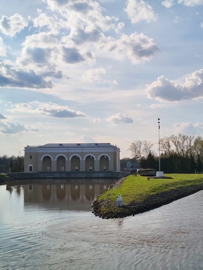 The image shows a building with a series of arched windows reflected in the water of a canal or river. The building is light yellow with a white trim. The water appears to be brown. To the right of the building is a grassy area with a flagpole. A white navigational marker sits on the edge of the grassy area. The sky above is blue with puffy white clouds. The image appears to be taken near Водник Снт, Russia.