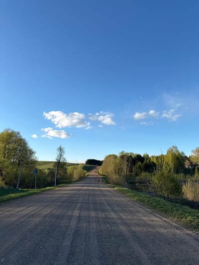 The image shows a rural dirt road under a clear blue sky with scattered cumulus clouds. The road is flanked by trees and green vegetation. There are utility poles visible along the roadside. The image does not provide enough detail to confirm or deny that this is located in Batmany, Russia.