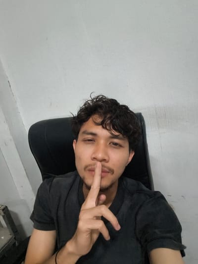 A young man with curly dark hair and a mustache is sitting in a black office chair. He is wearing a black t-shirt and has his index finger to his lips, in a "shushing" gesture. The background is a plain, light-colored wall with some subtle vertical markings. To the left of the man, a dark object, possibly a piece of furniture or equipment, is partially visible. The lighting appears to be indoor, and the overall mood is casual and possibly playful due to the gesture. The image is a selfie, taken from a close-up perspective focusing on the man's face and upper body. No text is visible in the image. The image was taken in Pulodolang, Indonesia.