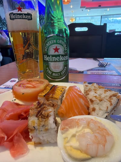 The image captures a meal served on a wooden table within a restaurant in Audun-le-Tiche, France.
In the foreground, a white, circular plate holds a variety of food items. These include a thick slice of red tomato, possibly resting on a white cheese base, accompanied by a pile of pink pickled ginger. Several sushi and maki pieces are present: one features a salmon fillet draped over rice, another shows a rectangular piece of yellow cooked egg (tamago) with a dark glaze over rice, and multiple rice rolls are topped with crispy fried onions and a light sauce drizzle. Additionally, a hard-boiled egg half is topped with a cooked shrimp and a dollop of white sauce, likely mayonnaise. A blue utensil handle is visible on the far right edge of the plate.
Behind the plate, a tall, clear glass branded "Heineken" is filled with a golden lager and topped with a white frothy head, exhibiting visible bubbles. Next to the glass stands a green glass bottle of "Heineken Original Pure Malt Lager," displaying condensation on its exterior.
The table beneath the items is made of light-brown wood, covered by a colorful paper placemat with fragmented text and graphics, including "AUDUN L..." and "AUTOSUP." A white paper napkin and a silver four-tined fork are visible on the placemat to the right of the drinks.
The background reveals a blurred restaurant interior with dark, possibly upholstered chairs featuring cut-out handles. Beyond the chairs, large windows show an indistinct exterior with cool-toned lighting. Overhead, a gold-colored chandelier and recessed lights illuminate the space. The ceiling is white with some red decorative elements. Blue and yellow ambient lighting contributes to the atmosphere. Partial, blurred figures of other patrons are visible in the far-left background, indicating an occupied establishment. No specific actions or interactions are discernible beyond the presentation of the meal.