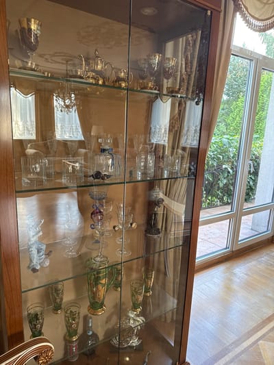 The image displays a glass-fronted display cabinet filled with various decorative items and glassware, positioned in a room with a wooden floor and large windows overlooking greenery. The cabinet is made of wood with glass shelves.
On the top shelf, there are gold-rimmed goblets and teacups, a gold teapot, a creamer, and a sugar bowl. A chandelier is visible reflected in the glass.
The second shelf from the top contains more glassware, including tall stemmed glasses and smaller cups. There is also a glass decanter with a blue ribbon.
The third shelf features several clear glass tumblers or whiskey glasses, some with metallic accents. A dark, possibly glass, object resembling a bird or animal is placed among them, and a small, decorative statue is visible on the right.
The fourth shelf holds a white ceramic figurine of a person playing a musical instrument, several clear stemmed glasses, and a green glass pitcher decorated with gold floral patterns. A dark glass bottle with a label is also present on this shelf.
The bottom shelf appears to contain more glassware and a silver decorative object.
The environment outside the window consists of lush green trees and foliage, suggesting an outdoor setting. The interior room has a warm, polished wooden floor and the overall impression is one of an elegantly furnished interior space. The location is identified as Yukki, Russia.