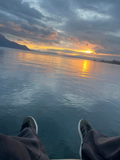 The image depicts a first-person, low-angle view over Lake Geneva (Lac Léman) in Montreux, Switzerland, during sunset. In the immediate foreground, the lower legs and feet of an individual are visible. The individual wears dark grey trousers and olive-grey sneakers featuring white soles and black stripes on the sides. Their feet extend horizontally over a narrow, light-colored surface, indicative of a boat deck or pier edge.
The vast expanse of Lake Geneva dominates the midground, presenting a calm, blue-green surface with minor ripples. A prominent vertical band of intense orange and yellow light reflects off the water, originating from the setting sun positioned low on the distant horizon to the right. The sky above is largely covered with dense, dark grey and blue-grey clouds, interspersed with areas of orange and subtle pink directly illuminated by the sunset. On the left side of the horizon, a dark mountain range, likely part of the Alps, is silhouetted against the colorful sky. To the right, along the distant shoreline, the silhouetted skyline of the city of Montreux is discernible. The overall scene is characterized by the distinct lighting of dusk.