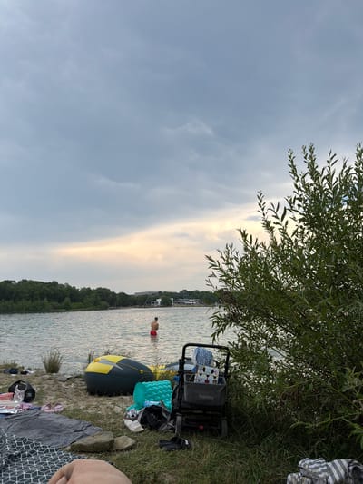 The image depicts a scene at a lake in Beckum, Germany, likely during an overcast day with a sliver of sunlight breaking through the clouds near the horizon. In the foreground, a person's leg is visible, suggesting the viewer is seated or lying down. To the left, a patterned blanket is spread on the grass, with various items scattered around it, including flip-flops, a JBL speaker, and a foil-wrapped object. A yellow and black inflatable boat rests on the shore.
Further into the scene, on the water, a lone individual is standing waist-deep, facing away from the camera. Across the lake, a tree-lined shore is visible, with some buildings and infrastructure on the far bank. To the right of the frame, a large, leafy green bush occupies a significant portion of the space, partially obscuring the background. A foldable cart with a fabric bag, containing items like a blue patterned cloth and what appear to be food packages, is positioned on the grass. The water is calm, with gentle ripples visible. The overall atmosphere is relaxed and suggests a day spent outdoors by the water.