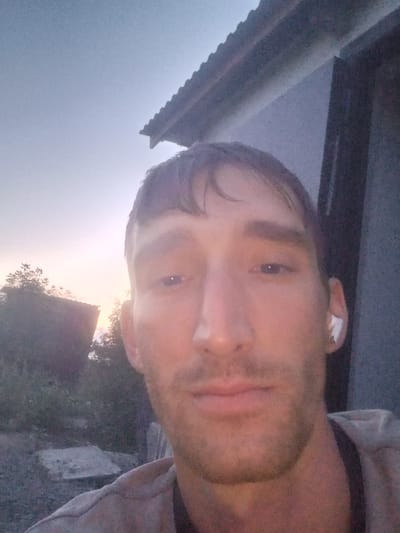 A medium shot selfie is taken from a slightly low angle by a fair-skinned man with short, dark hair and stubble. He is looking directly at the camera with a neutral expression. The man is wearing a light-colored t-shirt and has a white wireless earbud in his right ear. In the background, there is a partial view of a building with a corrugated roof. The setting appears to be outdoors, with trees and vegetation visible to the left, suggesting a rural or semi-rural environment. The sky in the background is a pale, dusky blue with a hint of pink, indicating either early morning or late evening. The location is identified as Қозыкеткен ауылдық округі, Kazakhstan. The overall lighting is dim, consistent with twilight hours.