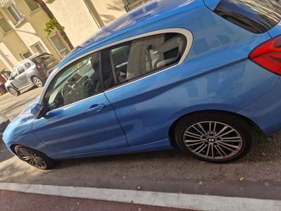 A blue BMW is parked on the street in Antony, France. The car is a hatchback and has a sunroof. The car is facing to the right of the image. The car is parked on a street with a sidewalk. There is another car, a silver Dacia Duster, parked behind the BMW. The buildings in the background are light yellow and have windows. The sky is blue and there are some trees. The sun is shining.