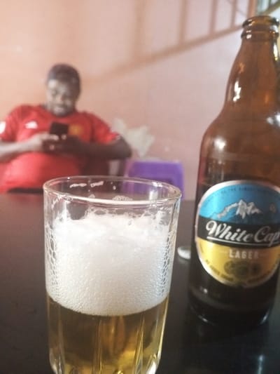 This image depicts an indoor scene located in Kisii Central ward, Kenya. In the immediate foreground, a clear glass filled approximately two-thirds with a golden-colored liquid, topped by a thick layer of white foam, rests on a dark, flat surface. To the right of the glass, a brown glass bottle, clearly identifiable by its label as "White Cap Lager," stands on the same surface. The bottle's label features a blue segment with a white mountain motif, and "White Cap" printed in white, with "LAGER" in gold.
In the blurred background, a dark-skinned individual, presumed male, is seated. He is wearing a red short-sleeved shirt and is actively engaged with a black smartphone, which he holds with both hands and looks down at. The background wall behind the individual is a plain, light orange color. A small purple object, possibly a stool or container, is partially visible to the right of the individual and behind the beer bottle. A vague white object is also discernible in the left background, near the individual's left side. A light-colored, possibly metallic or wooden, structural element is visible in the upper right portion of the background. There is no observed interaction between the visible subjects or objects beyond their arrangement within the scene.