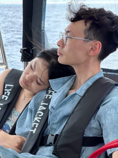 Two individuals are captured in a close-up shot, suggesting they are inside a boat due to the presence of water visible in the background. The male on the top wears a light blue button-down shirt, glasses, and a black life jacket. The female below wears a light blue button-down shirt and a black life jacket. "LIFE JACKET" is printed in white on the life jackets. The setting suggests a boat trip or water activity, possibly in Gulhi, Maldives, considering the context. They are looking down, and the boat appears to be in motion.