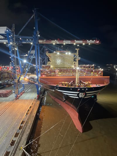 A nighttime aerial view captures a large container ship docked at the Port of Abidjan, Ivory Coast. The ship, branded with "MSC" on its bow, is positioned alongside a quay. A massive blue gantry crane, labeled "P5 KALMAR," extends over the ship and the port's paved surface, which is illuminated by numerous lights. The port area is filled with stacked shipping containers, some of which are visible on the left side of the frame. The water surrounding the ship appears muddy. The ship's hull is dark red, with white and black stripes on the bow. The upper decks of the ship are stacked with containers, and the text "NO SMOKING" is visible on the superstructure. The overall scene depicts a busy commercial port operating during nighttime hours.