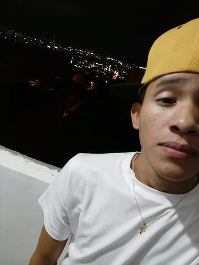 A young man with tan skin, wearing a white t-shirt and a mustard-yellow baseball cap, poses for a selfie at night. He has a gold chain with a cross pendant. In the background, a dark landscape is dotted with sparkling city lights. A white ledge is visible to the left of the frame. The man looks directly at the camera, creating a casual yet confident portrait.