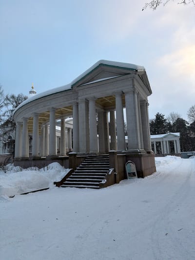 The image displays a snow-covered neoclassical colonnade in посёлок дачного хозяйства «Архангельское», Russia, under an overcast light blue sky, brighter towards the upper right. The central subject is a large, light-colored curved colonnade, featuring numerous columns supporting a roof, with a straight end section topped by a pediment on the right. A thick layer of white snow covers the roof, revealing a green trim along its edge. Dark-colored steps, partially obscured by snow, ascend to the raised base of the colonnade. The ground is uniformly covered in snow, with significant drifts accumulated to the left of the stairs. A snow-cleared path extends to the right, adjacent to which stands a light-colored A-frame sign with dark lettering and a logo. In the background, a white-domed building with a golden, cross-topped spire is visible behind the left curve of the colonnade, and another smaller columnar structure is present further right amidst snow-dusted bare and coniferous trees. No individuals are present.
