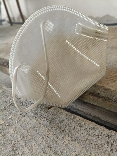 The media file is a close-up, eye-level shot of a white KN95 mask resting on a rough, weathered wooden surface. The mask is positioned at an angle, with its elastic ear loops visible. The texture of the mask, with its woven pattern and sealed edges, is clearly discernible. Sunlight casts highlights and shadows across the mask, suggesting it is daytime, likely outdoors due to the natural lighting and rustic setting. The background is blurred, showing hints of what appear to be industrial or construction elements, possibly indicating an outdoor workshop or storage area. The wooden surface itself is worn and appears to be part of some sort of bracing or scaffolding. There is no visible text in the image. The overall impression is one of a discarded or temporarily placed personal protective item in a utilitarian environment. The location context provided is Grinăuți, Moldova.