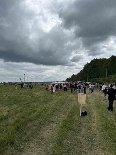 The image depicts an outdoor event with a crowd of people gathered in a grassy field under a cloudy sky. In the background, a dense line of trees forms a dark green border along the horizon. Several tents and stalls are visible among the crowd, suggesting a market or festival. People are seen walking, standing, and interacting. The ground is covered in green grass, with some areas appearing dry and sparse. There are also a few scattered objects, including a wooden signpost with a blank rectangular panel supported by a single wooden post and a black bag at its base. In the mid-ground, a tall, slender structure resembling a crane or gantry is present. The overall atmosphere suggests a daytime gathering, possibly in the city of Nurkeevo, Russia, given the context provided.