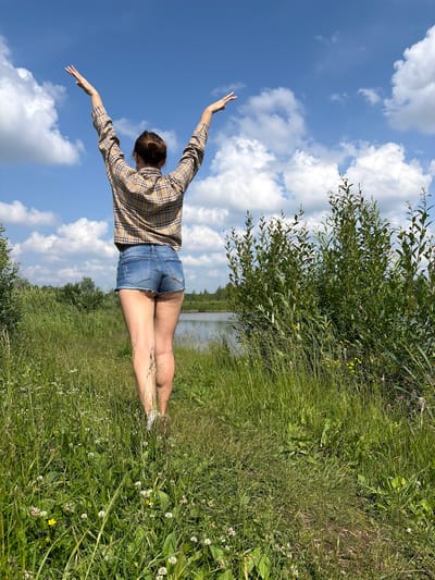 A woman stands in a grassy field with her arms raised to the sky. She is wearing a plaid shirt and denim shorts. The field appears to be near a body of water and is surrounded by trees. The sky is blue with scattered clouds. This scene is not located in Gushchino, Russia.