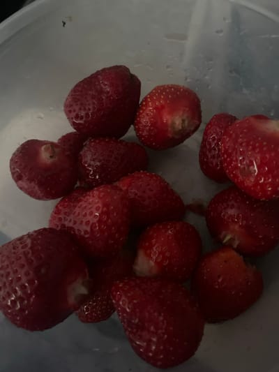 The image shows a close-up, overhead view of a collection of strawberries in a clear plastic container. The strawberries are mostly whole, with a few showing signs of having been bitten or cut. Their color ranges from bright red to a deeper, darker red. The container's surface is slightly wet, with droplets of water visible. The background is dark and out of focus, with some bright white reflections. The location in Sosnovka, Russia is not discernible from the image itself.