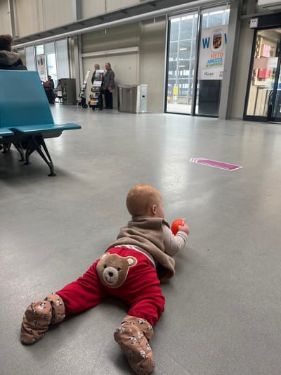 The image depicts an indoor scene within an airport terminal located in Lautzenhausen, Germany. The primary subject is an infant lying prone on a light gray floor in the foreground, facing towards the right. The infant wears red pants with a brown teddy bear face design on the rear, a light-colored long-sleeved top, a brown sleeveless vest, and patterned light brown booties. Its right hand grasps a small, orange, cylindrical object.
In the midground to the left, a section of teal-colored multiple-person airport seating is visible. Further in the background, to the left of center, two adults stand near a multi-tiered display stand holding brochures or magazines; one appears to be an older female with light hair, and the other an older male in a dark jacket. A stainless steel trash receptacle is positioned to their right. Beyond these elements, large glass doors and windows open to an exterior view, revealing what appears to be part of an airfield or parking structure. A prominent advertising poster affixed to one of the glass panels reads "FLY TO ABRUZZO, BE SURPRISED!" with airline branding including "Alitalia". A purple rectangular floor marking is visible on the light gray floor, further towards the right background. Other indistinct figures are partially visible in the distant background.