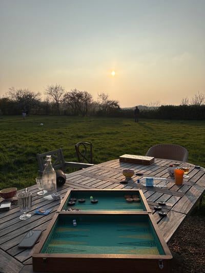 The image depicts an outdoor recreational scene at sunset in Bû, France. In the foreground, a rectangular wooden slatted table is positioned on a gravel surface. An open backgammon board, featuring a green felt playing surface with black and brown checkers, and two white dice showing "2" and "4," occupies the center of the table. To the left of the backgammon board are a clear glass bottle, two empty drinking glasses, a red and white patterned bowl, a silver lighter, a smartphone, and a small pack of cards. To the right, a clear plastic water bottle lies horizontally, alongside an orange cup, a yellow and brown patterned bowl, and a closed rectangular wooden box. A wicker chair is partially visible to the right of the table, and two wooden chairs are positioned behind the table, with one appearing partially overturned.
Beyond the table, a vibrant green grassy lawn extends towards a line of trees and bushes forming a distant tree line. On the left side of the lawn, a small figure, likely a child, stands near a white soccer ball. Further to the right, another individual stands facing away from the viewer, appearing as a dark silhouette against the bright sky. The sky above is a pale orange and yellow hue, indicating late afternoon or early evening, with a distinct, bright yellow sun positioned low on the right side of the horizon. The overall environment suggests a rural or semi-rural setting outside a residential structure, characterized by leisure activities under a sunset sky.