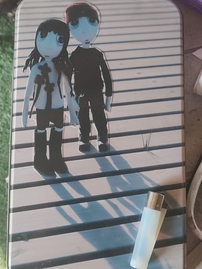 The image depicts two stylized, doll-like figures standing on a slatted wooden surface. The figure on the left has long black hair, a white top with black accents, dark shorts, and black boots. The figure on the right wears a black long-sleeved shirt and dark pants, with a red beanie. Their facial features are large and bulbous, with dark eyes and prominent red lips. The overall aesthetic is stark and somewhat somber. A pale, translucent object, possibly a lighter, lies at an angle on the wooden surface in the lower right quadrant of the image. The background is blurred, with hints of green suggesting an outdoor setting, and the lighting creates distinct shadows cast by the figures. The scene is likely located in Santa Lucía de Tirajana, Spain.