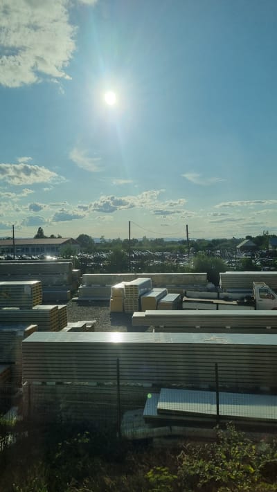 This is an exterior, medium-shot photograph of a construction material storage yard on a bright, sunny day. The sky is a clear blue with scattered white clouds, and the sun is high and shining brightly, causing lens flare. In the foreground, stacks of pre-fabricated construction panels, likely insulated metal panels, are neatly arranged. These panels are silver and have a ribbed texture. Some of the stacks are wrapped in plastic. Behind the panels, a gravel-covered area is visible, with more stacks of panels and other construction materials. Further back, there is a long building with a red roof, possibly a warehouse or administrative building. Trees and utility poles are scattered throughout the landscape. In the distance, a hazy horizon line suggests rolling hills or fields. The overall impression is one of an organized industrial or commercial space, likely a supplier of building materials. The location is identified as Fântâna Mare, Romania.