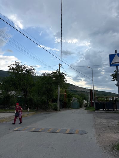 The image depicts a street in Zaprudnoe, Ukraine, oriented in an oblique perspective. A woman in a red headscarf and patterned clothing is walking on the right side of the street, next to a speed bump. The street surface is asphalt with visible wear. Along the sides are vegetation, fences, and sporadic structures. Power lines run overhead. In the distance, a hill is visible. The sky is filled with clouds, indicating potential overcast weather. A road sign is seen in the upper left corner.