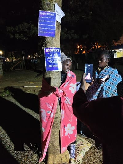In a dark outdoor setting, a woman in a knit hat stands near a tree, wrapped in a pink blanket. Multiple signs are attached to the tree, one promoting "Single Mothers" and another advocating for "Respect To all Single Mothers," citing "Hugging tree with single mothers benefits the society" for "68 Hrs." Another woman uses a smartphone to photograph the first woman, while a third woman stands by, looking at a small phone.