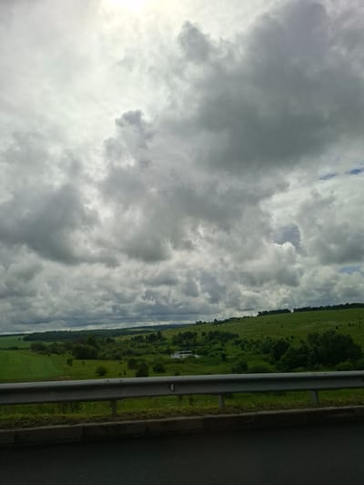The image shows a view of a rural landscape under a heavily overcast sky, likely in or around Lesnye Morkvashi, Russia. In the foreground, a gray metal guardrail runs horizontally across the lower portion of the frame, separated from the dark asphalt road by a strip of grass and a concrete curb. Beyond the guardrail, a green field stretches out, with rows of crops visible on the left. Further in the distance, rolling hills covered in lush green vegetation and scattered trees rise and fall. A small body of water, possibly a river or pond, is visible in the mid-ground, nestled amongst the trees. The sky is dominated by large, dramatic grey clouds, with patches of brighter sky peeking through in some areas, suggesting either the early stages of a storm or the clearing after one. No people or animals are visible in the scene.