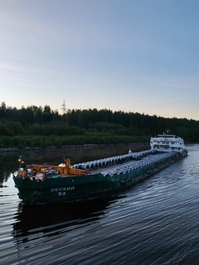 This image captures a long cargo barge, identified as "ОКСКИЙ 53" (OKSKIY 53), navigating a tranquil river or canal in the Depo region of Russia. The barge is laden with a substantial cargo of large, cylindrical rolls, likely steel coils, neatly arranged in rows across its open deck.
The bow of the vessel, painted dark green, prominently displays the ship's name "ОКСКИЙ" and number "53" in white Cyrillic letters. This area features an orange-yellow deck structure with various mechanical components and a small flag. At the stern, a multi-story white superstructure, presumably the bridge, is visible, also bearing Cyrillic text that appears to read "ЖКСНИ И Д2" (ZHKSNI I D2) and the number "53".
The waterway is bordered by a lush, dense forest of dark green trees, whose reflections are subtly visible on the calm water. In the background, a large metal power line pylon rises above the treeline, with power lines stretching across the upper portion of the sky. The time of day appears to be either dawn or dusk, as suggested by the soft, ambient light and the sky's gentle gradient from light blue at the zenith to a pale yellow closer to the horizon. A slight wake trails behind the moving barge, indicating its forward momentum. No people are visible. The scene conveys a sense of peaceful industry within a natural landscape.