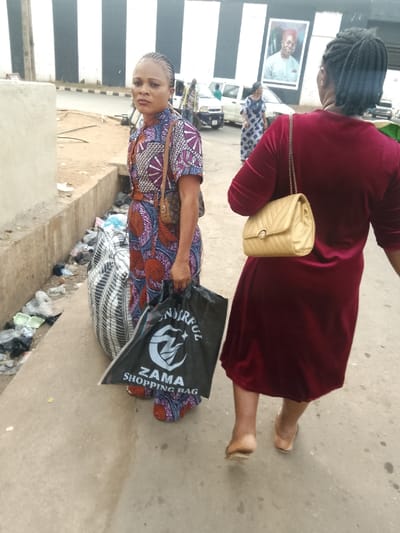 Two women walk along a street with trash strewn nearby. The woman on the left faces forward, wearing a colorful patterned dress, carrying a handbag, and a black โZama Shopping Bag.โ Beside her, a large striped bag rests on the ground. The second woman, seen from behind, wears a deep red velvet dress and carries a light-colored purse. Further in the background, other figures and vehicles are visible under an overpass with a billboard.