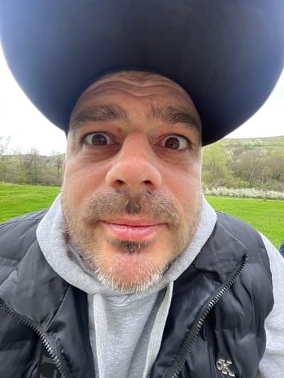 A selfie captures a man in Barkach, Bulgaria. He wears a black baseball cap and a black puffer vest over a grey hooded sweatshirt. The background features a grassy field with trees.