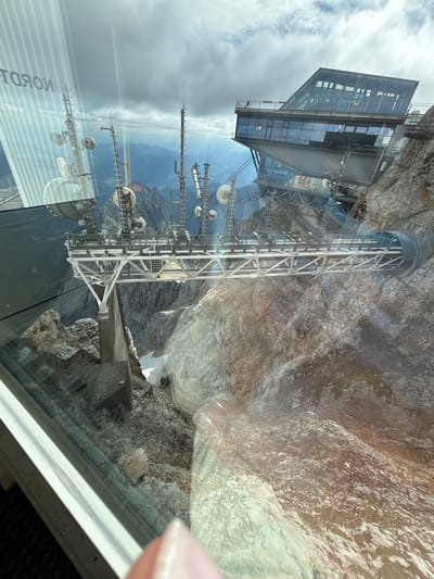 The image shows a view from a window looking out at a mountainous landscape. In the foreground, a modern glass building with angular lines is perched on the side of a rocky mountain. A metal structure, possibly a walkway or part of a communication tower, extends from the building over a steep drop. Various antennas and satellite dishes are mounted on this structure. The background reveals a panoramic view of rugged, grey mountains under a cloudy sky. The reflection on the glass in the foreground shows vertical stripes and what appears to be text, possibly from an advertisement or sign. The overall scene suggests a location at a high altitude, likely a tourist attraction or a research facility in a mountainous region. The image is taken from inside a moving vehicle, likely a cable car, given the slight blur and the perspective. The mention of Ehrwald, Austria, suggests this location could be a prominent peak or viewpoint accessible from the town.