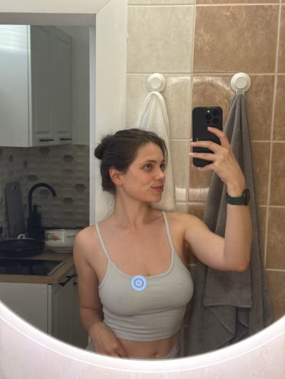 A woman is taking a selfie in a bathroom mirror. She is wearing a gray tank top and a green smartwatch. She is holding a black smartphone in her right hand. The bathroom has beige and white tiles. There are two towels hanging on the wall, one gray and one white. A white cabinet and a kitchen sink faucet are visible in the reflection. The mirror is round with a white frame and a glowing power button. The location is a bathroom, presumably in Kőröshegy, Hungary.