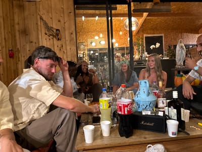 The image depicts an indoor gathering of approximately six to seven individuals seated around a wooden table. The setting appears to be a casual, perhaps domestic, environment with a rustic ambiance, featuring wooden paneled walls and ceiling. One wall has decorative elements, including what appears to be dried reeds and a cicada figurine. Large glass doors or windows reveal an illuminated interior space beyond, with more wooden paneling and hanging lights.

The individuals are diverse in gender and ethnicity, and their attire suggests a relaxed social event. Some are looking at each other, indicating conversation and interaction, while others are looking away or downward. A prominent feature on the table is a collection of bottles, including what appears to be Coca-Cola, Fanta, Captain Morgan Spiced Rum, and Ballantine's whisky, along with several white paper cups. A black Marshall speaker is also visible on the table, suggesting music may be playing. Other items on the table include sunglasses, a lighter, and what seems to be a pack of cigarettes.

The overall impression is of a social gathering with drinks and possibly smoking, taking place in what could be a communal space within a home or a rented venue. The lighting is warm and somewhat dim, contributing to the cozy atmosphere. The scene is devoid of any specific landmarks or clues that would precisely pinpoint the location to Solliès-Pont, France, without additional context.