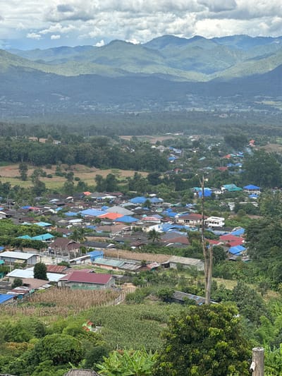 An elevated, panoramic view depicts the village of Ban Mai Saha Sampan, Thailand, nestled within a valley under an overcast sky. In the background, multiple layers of lush green, forested mountains extend across the horizon, gradually fading in hue due to atmospheric perspective. The midground features a wide expanse of dense forest and some cultivated fields. The foreground and lower midground are dominated by the village itself, a dense cluster of residential structures characterized by numerous multi-colored roofs, predominantly bright blue, light grey, and red, intermingled with dark green foliage. Patches of agricultural land, possibly cornfields, are visible between and around the houses. A prominent, vertical, light grey dead tree trunk stands in the right mid-foreground. No people or discernible actions are visible. The environment is rural, mountainous, and tropical.