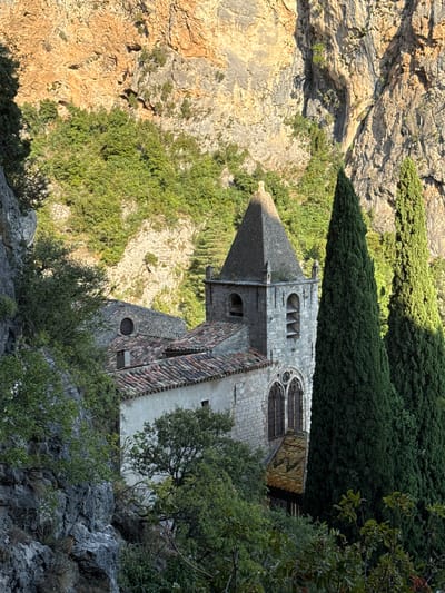 This image depicts a stone church with a tiled roof and a steeple, nestled within a rugged, rocky landscape covered in green foliage. The church is partially obscured by trees, with tall, slender cypress trees flanking the right side of the steeple. The background consists of a steep, rocky cliff face with patches of vegetation. The scene appears to be illuminated by natural light, casting shadows and highlighting the textures of the stone and rock. The architectural style of the church, with its arched windows and stone construction, suggests it is an old building. The overall impression is one of a remote, historical structure integrated into a natural, mountainous environment. The location is identified as Moustiers-Sainte-Marie, France, a village known for its dramatic setting within a gorge.