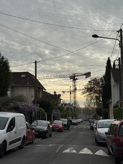 The image depicts a street scene in Champigny-sur-Marne, France. Cars are parked along the right side of the street, facing the same direction. The street narrows in the distance. A construction crane is visible in the middle distance, extending over the rooftops of buildings. Utility poles and wires run along the side of the street. The sky is overcast. A woman in a green dress can be seen walking along the street. Some foliage and flowering trees can be seen in the foreground.