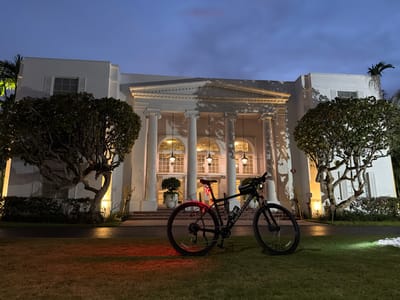 A black mountain bike is parked on a grassy lawn in front of a large, white building with a classical architectural style. The building features a grand entrance with several large white columns, arched windows, and ornate hanging lights. The scene is illuminated by artificial lighting, casting a warm glow on the building's facade and creating shadows from the trees. The sky above is a deep blue, indicating either dusk or dawn. A red taillight is illuminated on the bicycle. The location is identified as 10150 Collins Avenue in Bal Harbour Village, Florida, United States of America.