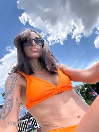 The image shows a low-angle selfie of a woman wearing an orange bikini. She has dark hair, sunglasses, and tattoos on her left arm. The background features a bright blue sky with white clouds. In the distance, there is a white structure, trees, and a Pepsi umbrella. The scene appears to be outdoors, likely in a sunny environment. No specific location in Grivitsa, Bulgaria can be identified from the image.