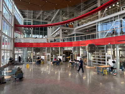 An expansive interior space with abundant natural light. People are seated at tables, engaged in various activities. A man in a mask walks across the polished floor. Above, red accents highlight the architectural structure. Multiple levels with glass partitions create visual depth. The space appears to be a modern academic or communal area. Unusual, spiky spherical art pieces punctuate the room.